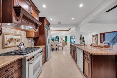 Bright open-concept luxury kitchen with dark wood cabinets, granite island and sink, stainless steel appliances and gas range, tiled floors, and a dining area framed by arched windows and lush tropical greenery.