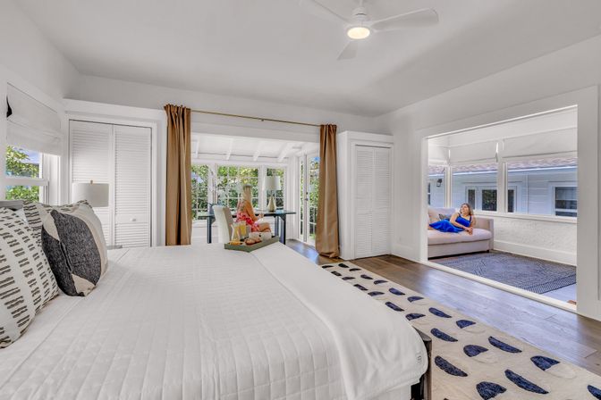 Sunlit coastal-style bedroom in a bright beach house with white quilted bed, patterned pillows, hardwood floors and rug, adjoining glass sunroom with desk and cozy sitting alcove.