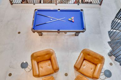Aerial view of a blue-felt pool table with crossed cues and racked balls in a modern indoor game room, flanked by two tan leather armchairs, round side tables, light tile floor and spiral staircase.