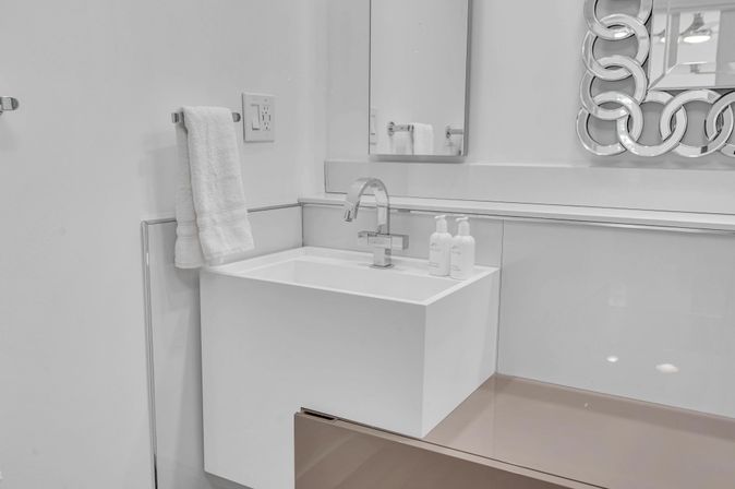 Modern white wall-mounted bathroom sink with chrome faucet, twin soap dispensers, hanging towel and decorative circular mirror