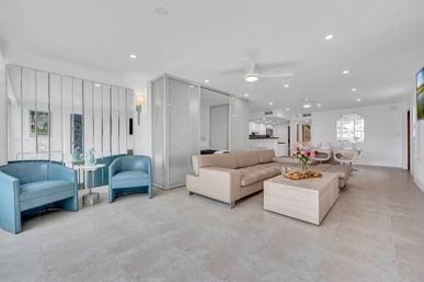 Bright, coastal-inspired open-concept living room with a beige leather sectional, blue accent chairs, mirrored wall, glass partition, large tile floors and a dining area in the background.