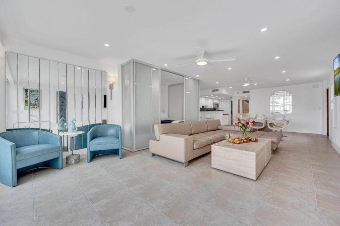 Bright, coastal-inspired open-concept living room with a beige leather sectional, blue accent chairs, mirrored wall, glass partition, large tile floors and a dining area in the background.