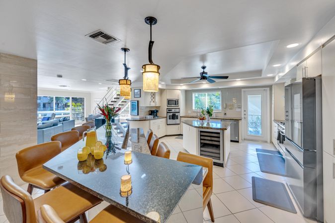 Modern open-plan kitchen and dining area with granite island and breakfast bar, leather stools, pendant lights, built-in wine fridge, stainless appliances, tile floor and bright windows overlooking an outdoor patio.