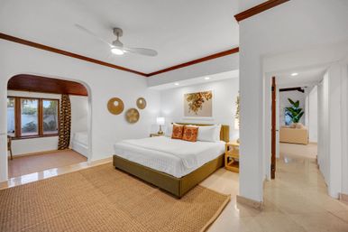 Bright, airy king bedroom in a vacation rental suite with white walls and wood trim, ceiling fan, woven jute rug, decorative wall baskets, archway to a windowed seating nook and open doorway to the living area.