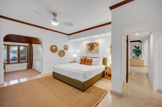Bright, airy king bedroom in a vacation rental suite with white walls and wood trim, ceiling fan, woven jute rug, decorative wall baskets, archway to a windowed seating nook and open doorway to the living area.