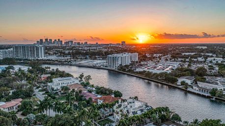 Aerial sunset view of a coastal city with a winding canal, palm-lined waterfront mansions, nearby luxury condos and a distant downtown skyline under an orange sky