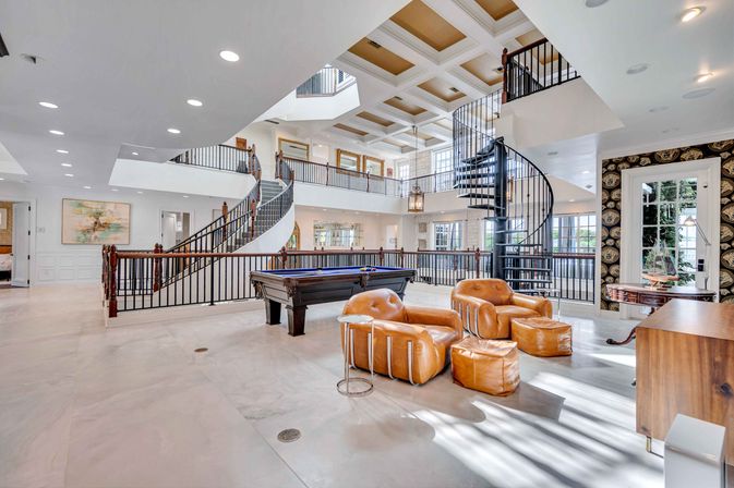 Sunlit luxury open-plan great room with a pool table, tan leather lounge chairs and ottomans, sweeping curved staircase, black spiral stair, coffered ceiling and bright mezzanine railings.
