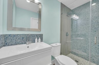 Bright modern bathroom with white vanity and sink, stainless-framed mirror, toilet, and glass-enclosed shower with blue mosaic tile and pebble floor, seafoam-green walls for a coastal vibe.