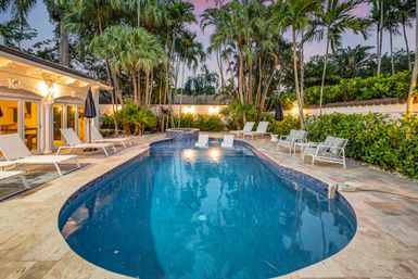 Tropical backyard pool at dusk with a curved blue swimming pool and raised spa, surrounded by palm trees, lounge chairs, umbrellas and a stone patio for outdoor relaxation