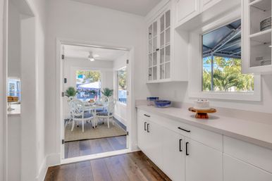 Sunlit coastal kitchen pantry with white cabinets and countertop, cake on a wooden stand, pass-through window and a bright breakfast nook with a round white table, blue-patterned chairs, and palm trees outside.