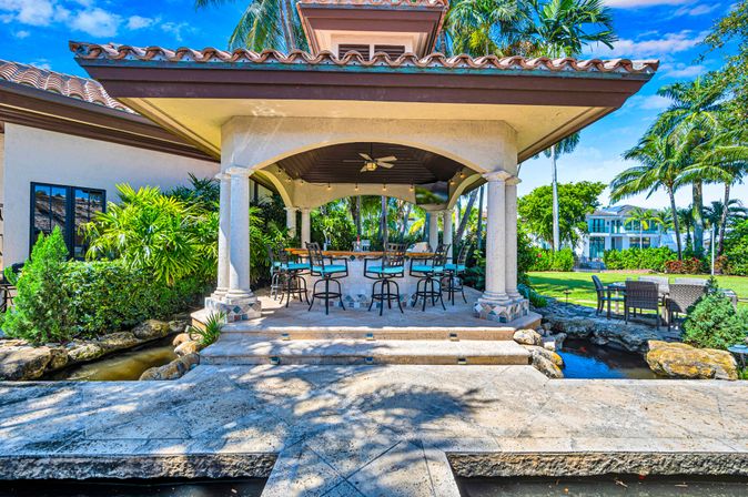 Sunlit Mediterranean-style waterfront patio with tiled roof and stone columns, outdoor bar with turquoise-cushioned stools, ceiling fan and string lights, surrounded by tropical palms, manicured lawn and canal.