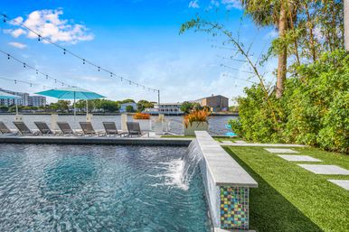 Riverside infinity pool with cascading edge, mosaic-tiled wall, row of sun loungers, turquoise umbrella, string lights and tropical landscaping