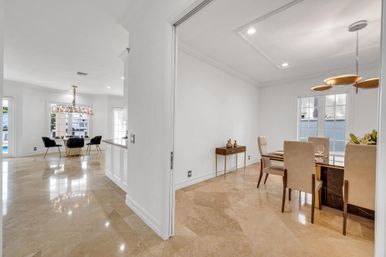 Sunlit open-concept dining area with polished beige marble floors, white walls, gold-accent chandeliers, modern table with beige upholstered chairs and large windows.