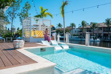 Sunny tropical waterfront pool and dock with a white cabana and yellow-striped cushions, in-pool loungers, palm trees, moored boats, and two people chatting on the wooden deck.