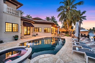 Luxury waterfront villa at sunset with curved pool and spa, palm trees, string lights, sun loungers on stone patio and a yacht at the private dock.