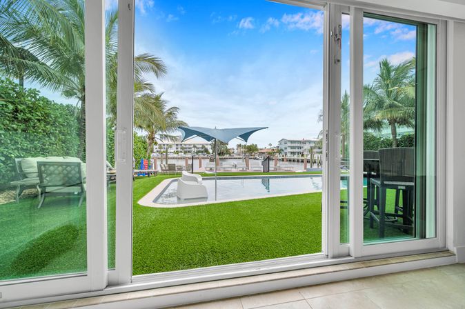 Sliding glass doors opening to a coastal poolside patio with bright green lawn, lounge chairs, a blue shade sail, palm trees and marina docks visible beyond