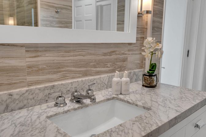 Sleek marble bathroom vanity with undermount sink, chrome faucet, matching pump bottles and a potted white orchid beside a lit wall sconce under a framed mirror.