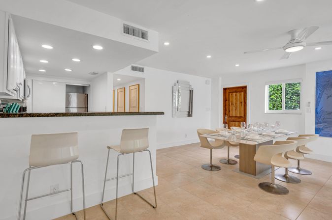 Bright modern open-plan kitchen and dining area with breakfast bar and two stools, dining table set for six with beige swivel chairs, tiled floor, wooden entry door, ceiling fan and a leafy window.