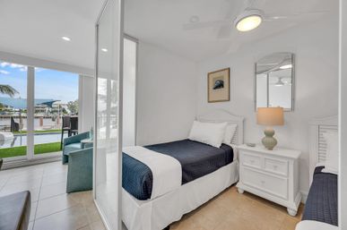 Bright coastal bedroom with two twin beds in navy quilts, white nightstand and lamp, sliding glass doors opening to a poolside patio and marina view with palm trees
