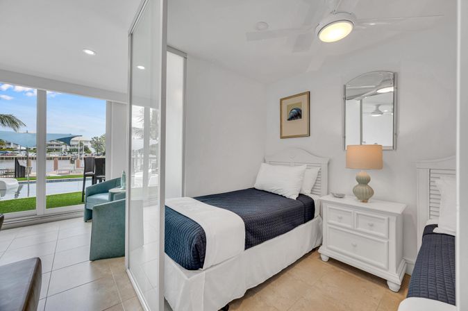 Bright coastal bedroom with two twin beds in navy quilts, white nightstand and lamp, sliding glass doors opening to a poolside patio and marina view with palm trees