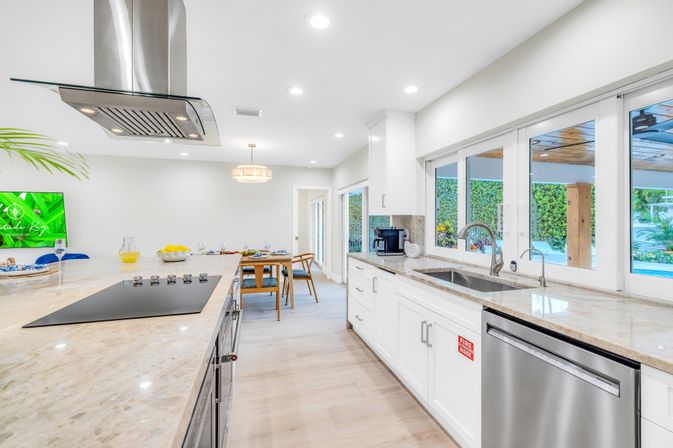 Bright modern open-concept kitchen with large marble island and black induction cooktop under a stainless range hood, white cabinetry, undermount sink beneath sliding windows overlooking a pool and tropical hedges, stainless dishwasher and a wood dining table in the background.