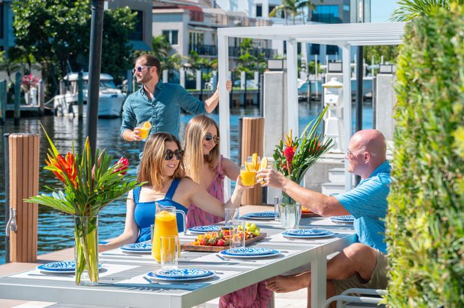 Group of four toasting with orange cocktails at a sunny waterfront dockside brunch by a marina, white outdoor table set with blue patterned plates, pitcher of orange juice and tropical flower centerpieces.