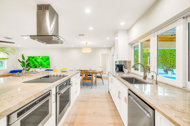 Sunlit open-concept modern kitchen with a large marble island, stainless steel range hood and appliances, undermount sink, sliding glass doors revealing a backyard pool, light wood floors and a cozy dining area.