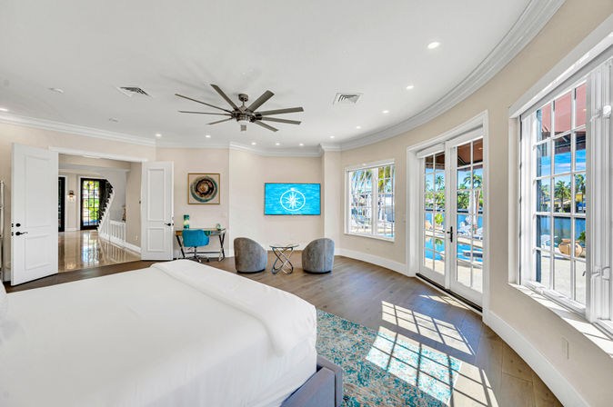 Sunlit waterfront master bedroom with king bed, large ceiling fan, seating area and desk, curved wall of windows and French doors opening to a pool and marina with palm trees.