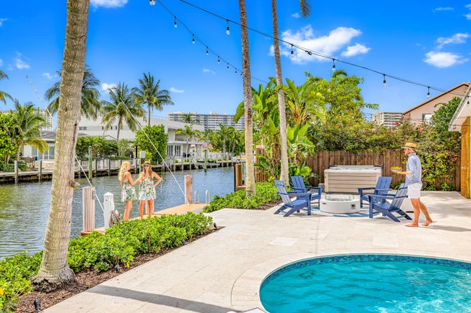 Sunny tropical waterfront backyard with round pool, patio chairs around a fire pit and hot tub, palm trees and string lights by a canal dock where two women chat and a man carries a tray.