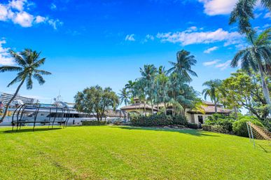 Sunlit waterfront tropical backyard with lush green lawn, palm trees, Mediterranean-style villa and a docked luxury yacht under a bright blue sky