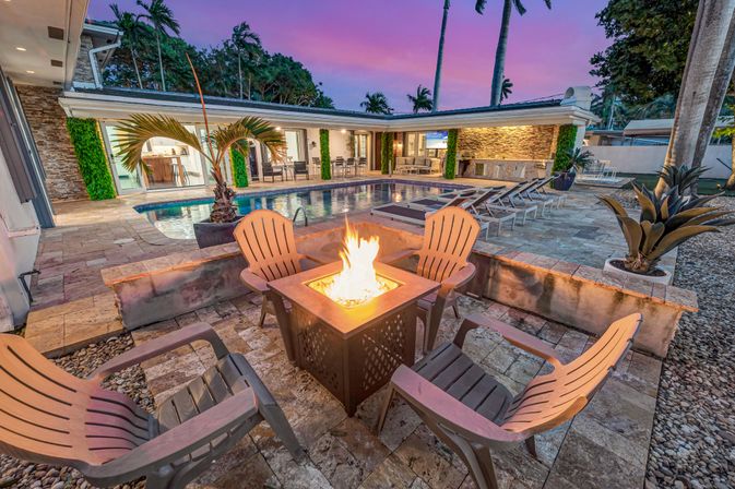 Luxury tropical backyard patio at dusk with a lit fire pit surrounded by Adirondack chairs, stone pool and lounge chairs beside a modern open-plan home with palm trees