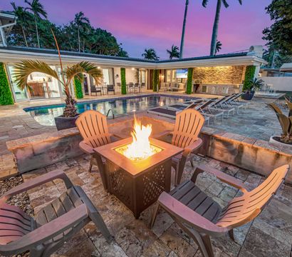 Luxury tropical backyard patio at dusk with a lit fire pit surrounded by Adirondack chairs, stone pool and lounge chairs beside a modern open-plan home with palm trees
