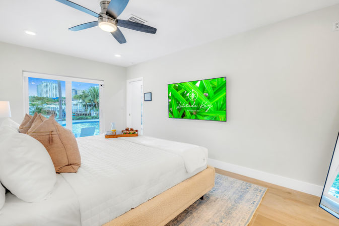 Bright modern waterfront bedroom with white linens and tan throw pillows, ceiling fan, wall-mounted TV, breakfast tray on the bed, and sliding glass doors opening to a pool and palm trees.