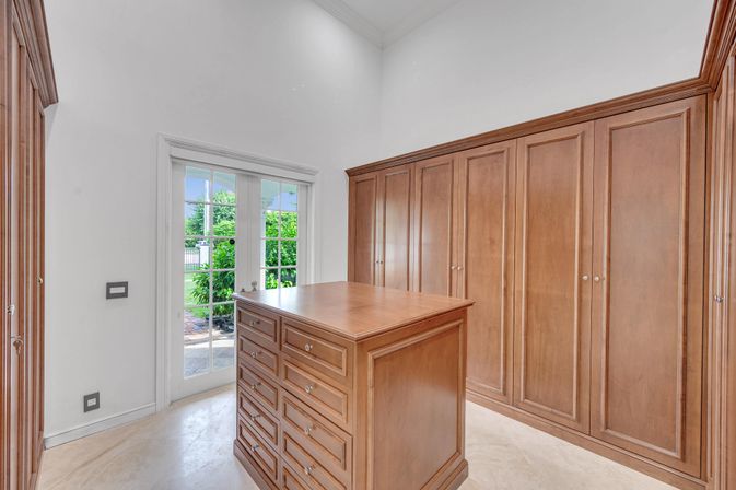 Sunlit residential walk-in closet with floor-to-ceiling wood cabinets, a central island of drawers, and glass French doors opening to a lush garden patio.