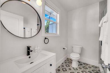 Bright modern white bathroom with round mirror, black faucet and hardware, vanity sink, window with blue sky view, toilet, and geometric patterned floor tiles — clean, airy design.