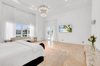 Luxury waterfront master bedroom in a coastal home — airy white interior with high ceilings and crystal chandelier, king bed with white linens, tufted sofa by floor-to-ceiling windows and patio doors opening to a canal view