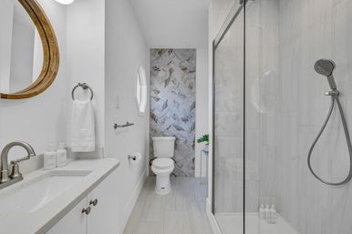 Bright modern white bathroom with a vanity and round wooden mirror, marble herringbone accent wall behind the toilet, glass walk-in shower with handheld showerhead, and chrome fixtures.