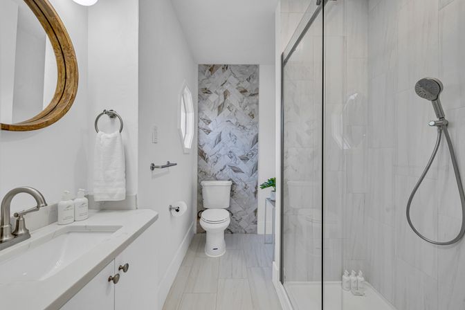 Bright modern white bathroom with a vanity and round wooden mirror, marble herringbone accent wall behind the toilet, glass walk-in shower with handheld showerhead, and chrome fixtures.