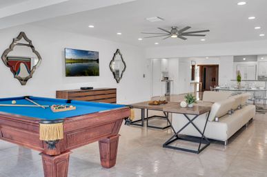 Bright open-concept living and game room interior with a blue-felt pool table, beige leather sofa, wall-mounted TV, decorative mirrors, modern ceiling fan and dining area on glossy tile floors