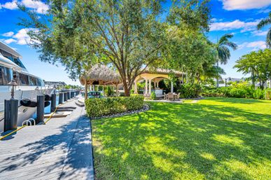 Sunny tropical waterfront backyard with wooden dock and docked yacht, lush green lawn, palm trees, thatched tiki hut and covered patio with outdoor seating