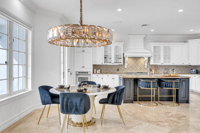 Bright open-concept luxury kitchen and dining area with marble floors, round white dining table surrounded by navy velvet chairs with gold legs, large crystal-and-gold chandelier, dark island with navy-and-gold barstools, tiled backsplash and stainless appliances, sunlit windows.