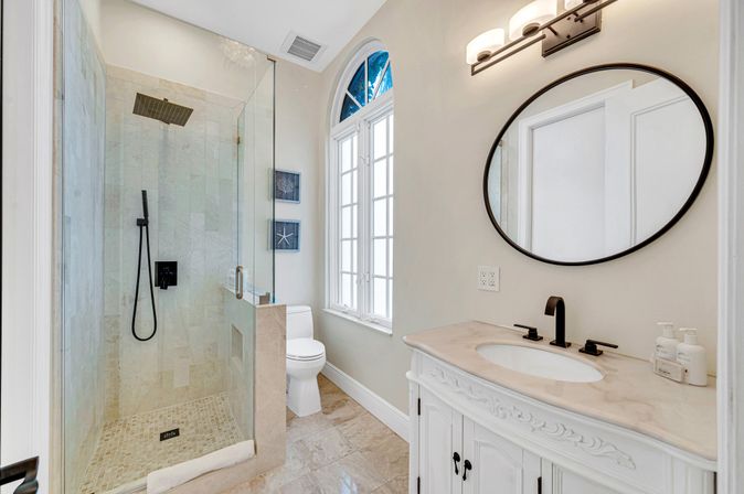 Spa-like luxury bathroom with glass walk-in shower and square rain showerhead, black matte fixtures, marble tile floor, white carved vanity with oval mirror, arched window and coastal wall art