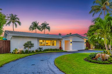 Single-story modern ranch home with two-car garage, curved asphalt driveway, lush tropical landscaping and tall palm trees, warm interior lights glowing beneath a vibrant sunset sky.