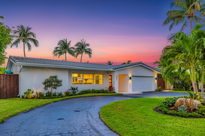 Single-story modern ranch home with two-car garage, curved asphalt driveway, lush tropical landscaping and tall palm trees, warm interior lights glowing beneath a vibrant sunset sky.