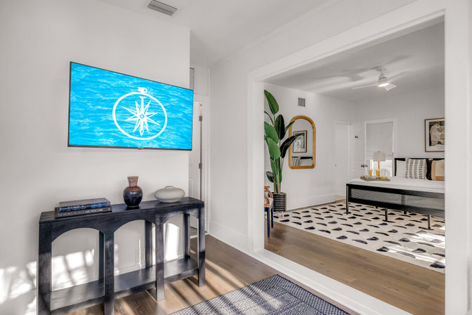 Sunlit modern coastal-style bedroom interior with hardwood floors; wall-mounted TV showing a blue compass graphic above a black console table with vases, open doorway to a bedroom with a patterned rug, potted plant, gold-framed mirror and minimalist bed.