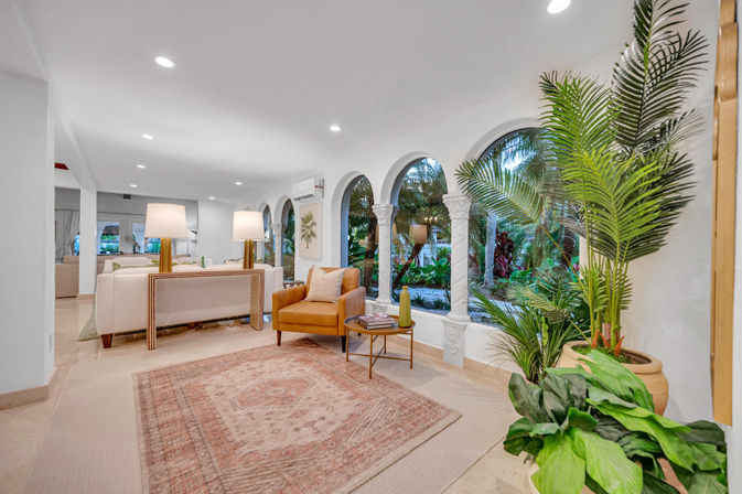 Inviting sunlit Mediterranean-style living room with arched windows overlooking a lush tropical garden, tan armchair, white sofa, brass table lamps, patterned rug and potted palms.