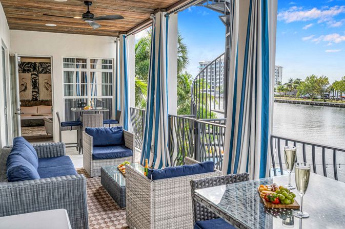 Canal-front tropical balcony with wicker seating and navy cushions, blue-and-white striped curtains, spiral staircase, glass table with champagne and a fruit platter, ceiling fan and palm-lined waterway view.