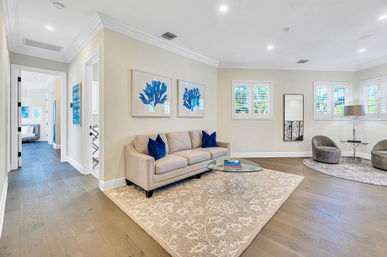 Bright, coastal-inspired open living room with a beige sofa and navy pillows, blue coral wall art, glass coffee table on a patterned rug, hardwood floors and sunlit windows with white shutters.