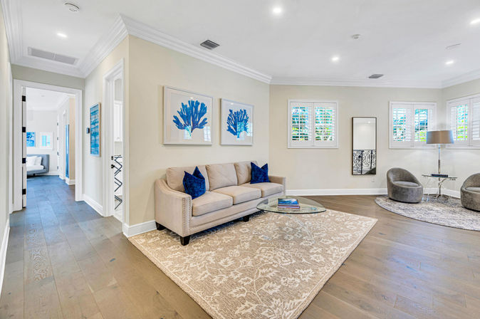 Bright, coastal-inspired open living room with a beige sofa and navy pillows, blue coral wall art, glass coffee table on a patterned rug, hardwood floors and sunlit windows with white shutters.