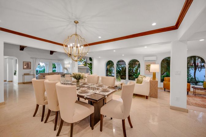 Bright open-plan dining and living room with beige upholstered chairs, modern gold chandelier, arched windows framing palm trees and a waterfront view